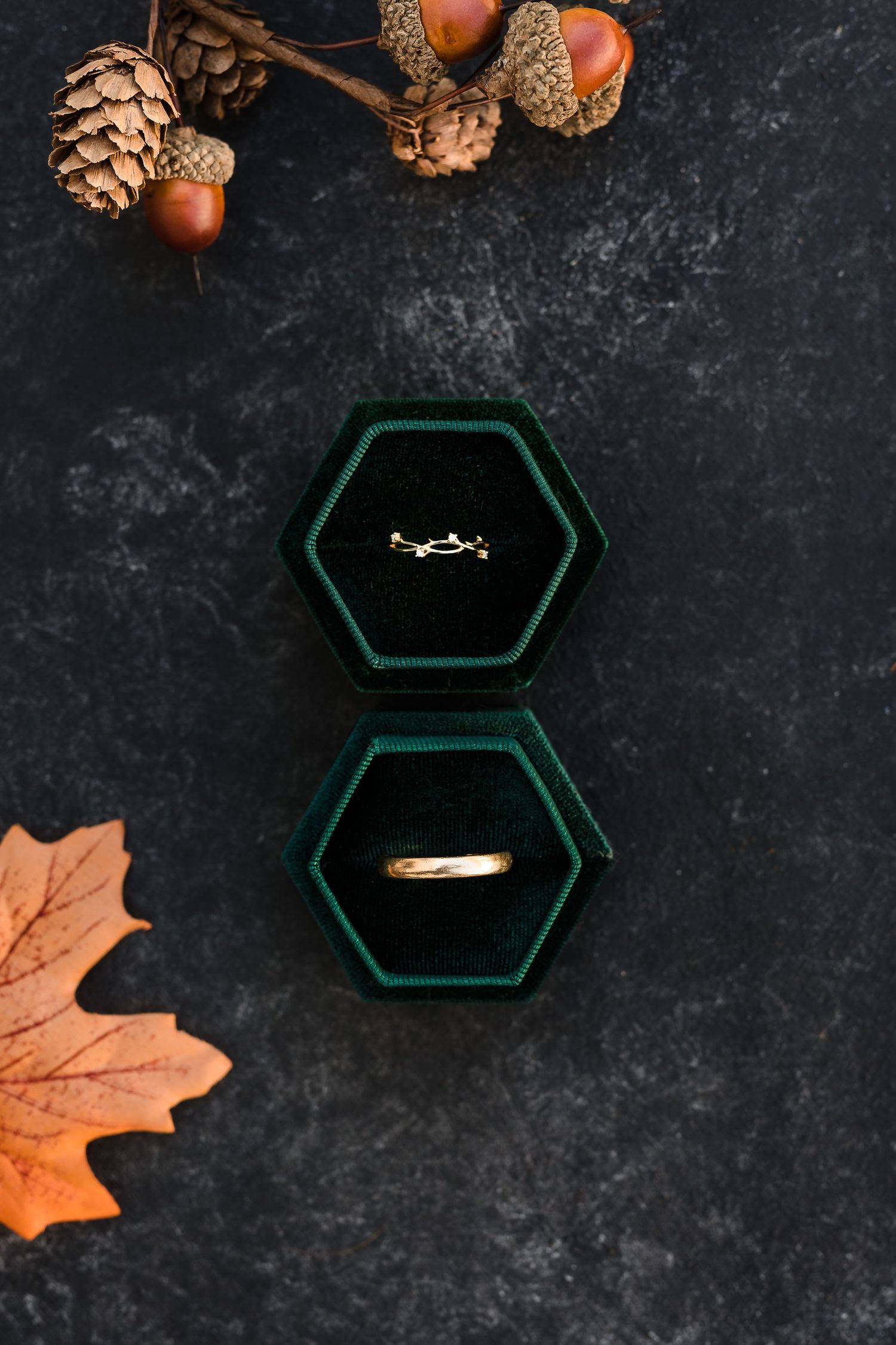 Two gold wedding rings in dark green hexagonal boxes with autumn pinecones and orange maple leaf on black surface.