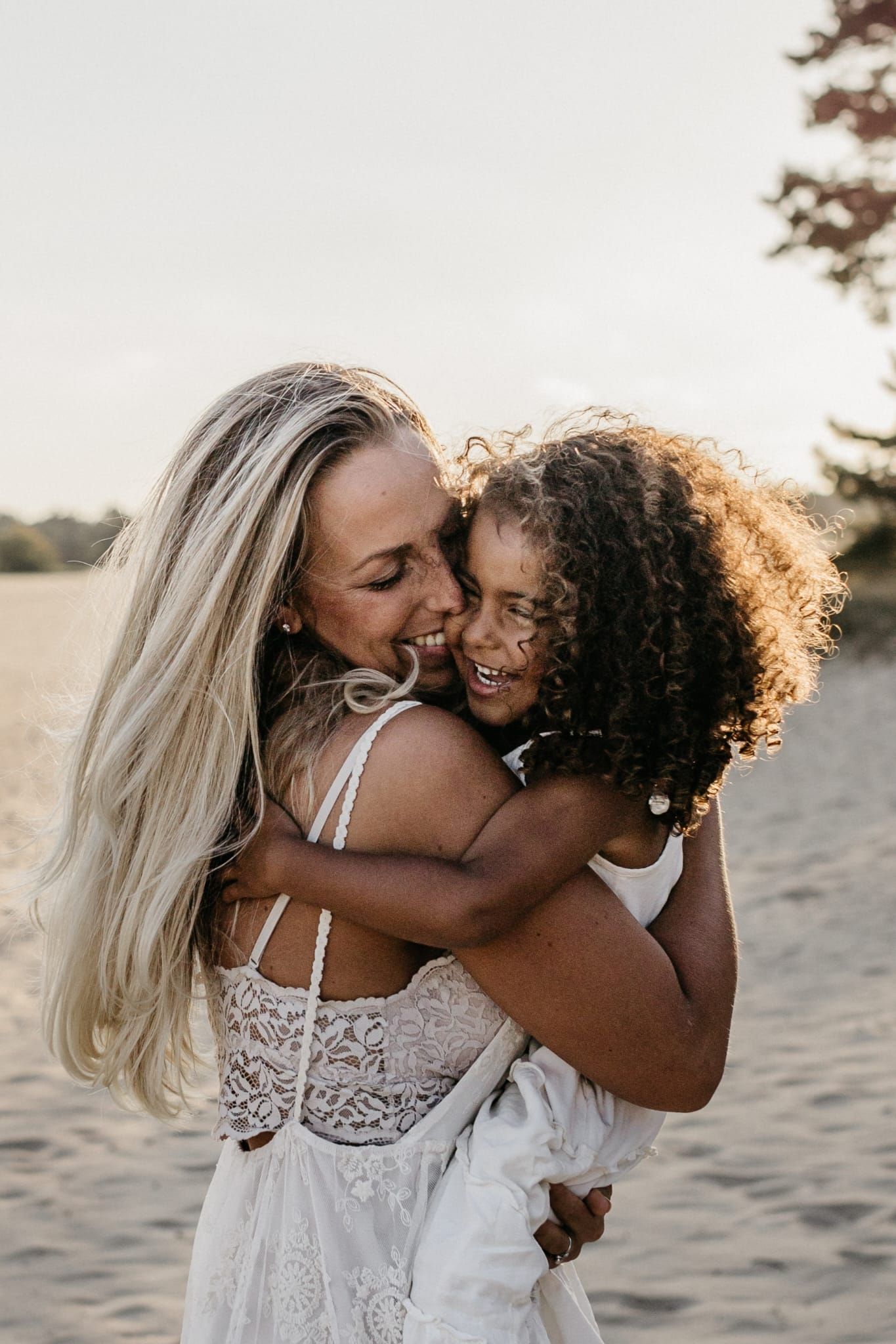 Joyful blonde mother hugging and kissing her curly-haired daughter on a sandy beach.