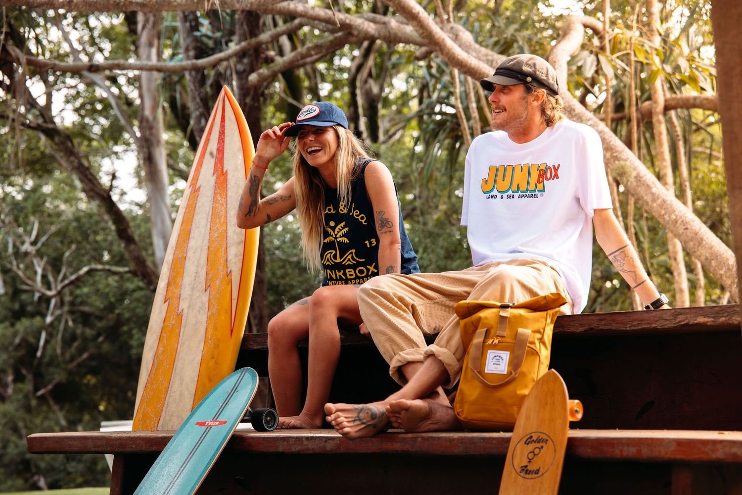 Two people relaxing with surfboards on wooden platform among trees.
