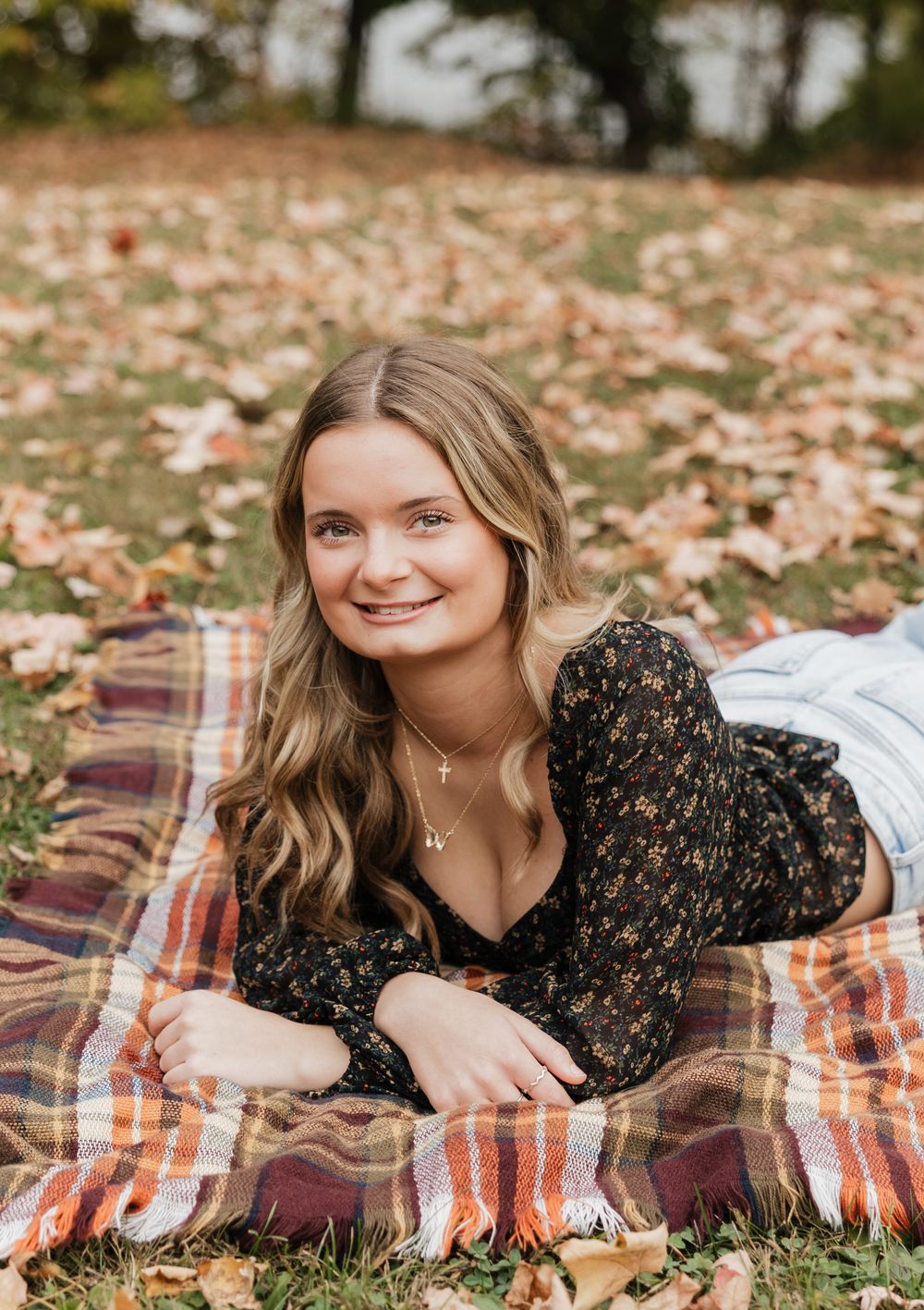 Person in black floral dress laying on plaid blanket surrounded by autumn leaves during outdoor photo session.