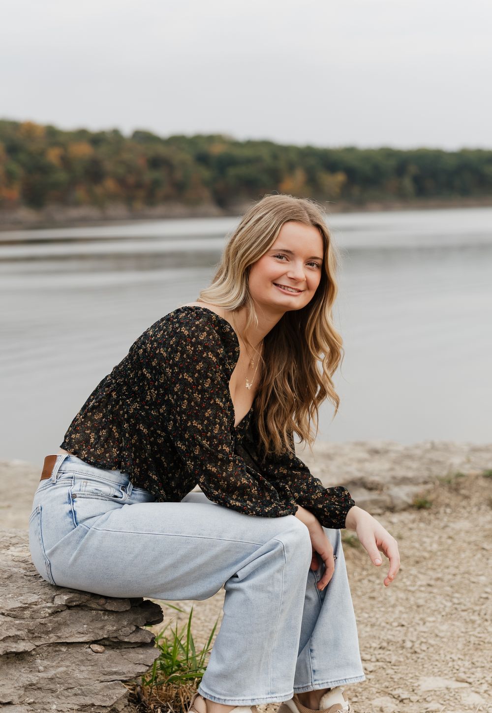 Someone in a floral top and light wash jeans sits casually by a lakeside on an autumn day.