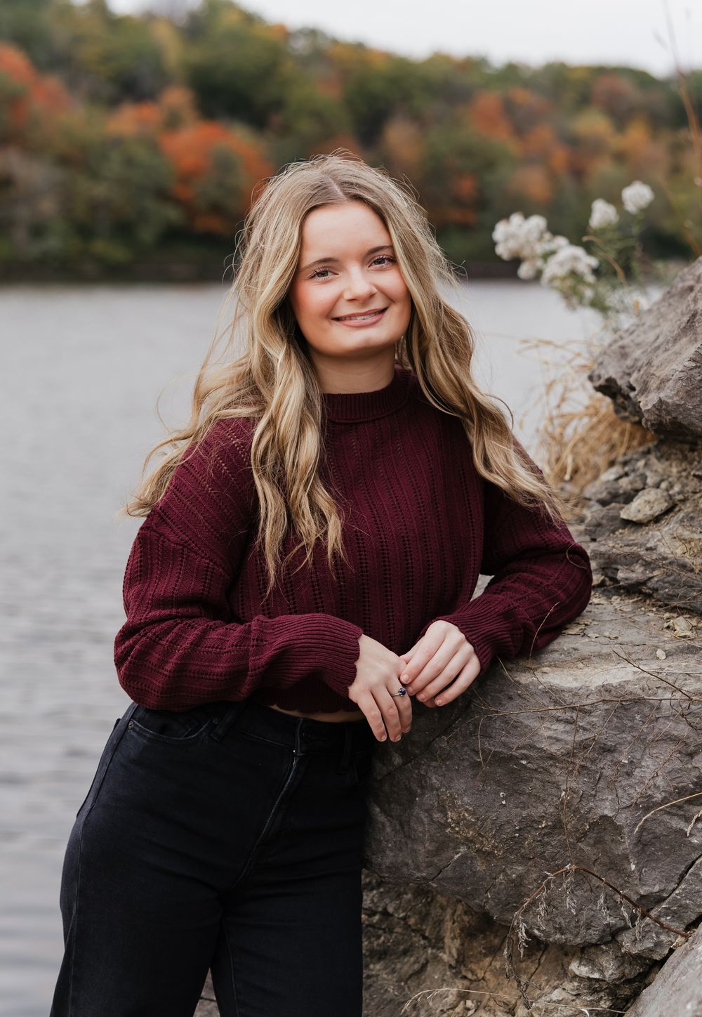 A person in a burgundy sweater poses by a rocky shoreline with autumn foliage in the background.
