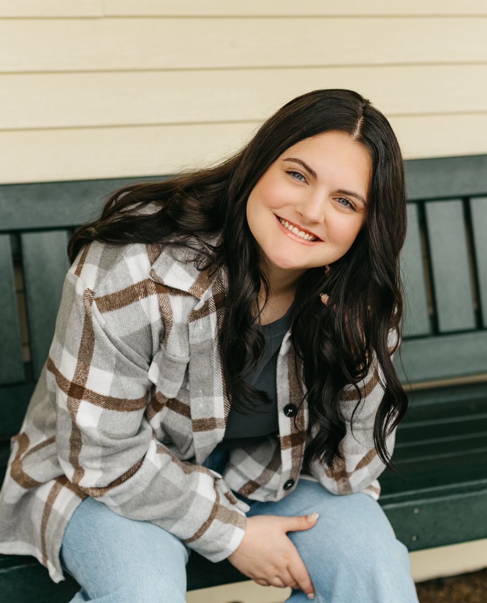 Someone sitting on a bench wearing a plaid flannel shirt and jeans with long dark hair and a bright smile.