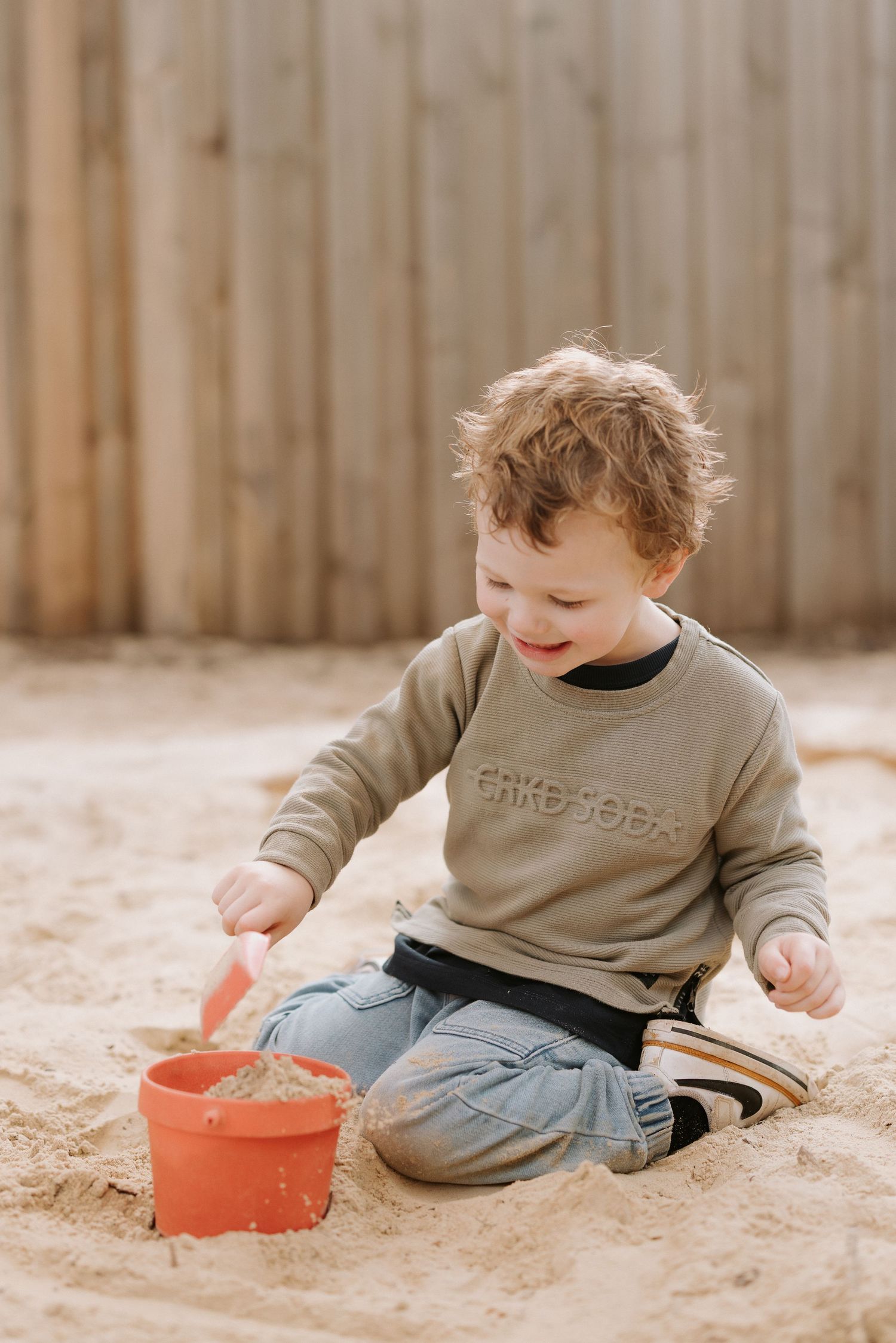 A toddler in a beige sweater and jeans plays with a red bucket in a sandbox on a sunny day.