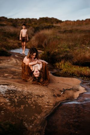 Mother and children enjoy a cuddly moment on rocky terrain near a stream at sunset.