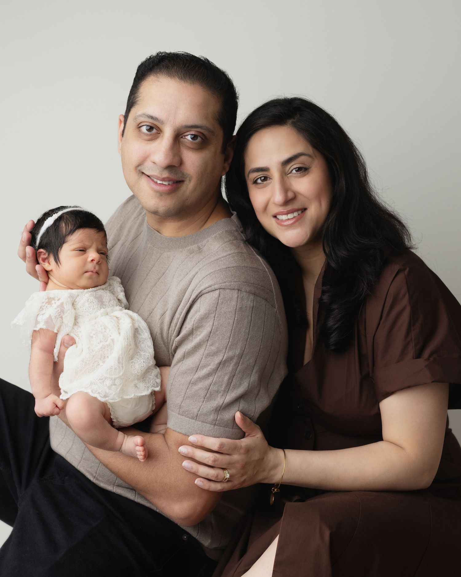 Family of three poses together in studio wearing brown and beige tones against light neutral background. Elizabeth Lang Photography Surrey