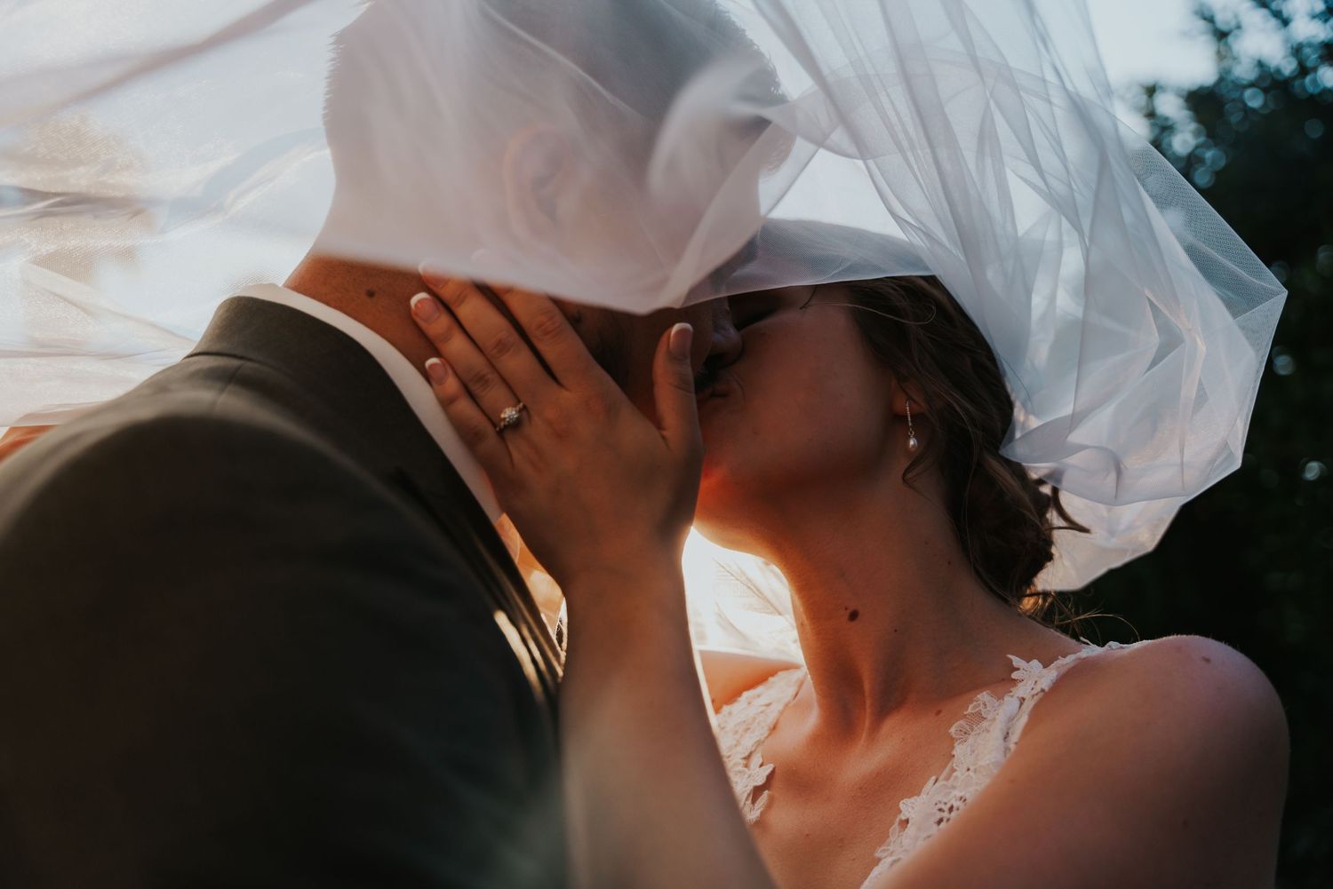 A couple shares a romantic kiss under a veil during a wedding ceremony outdoors.