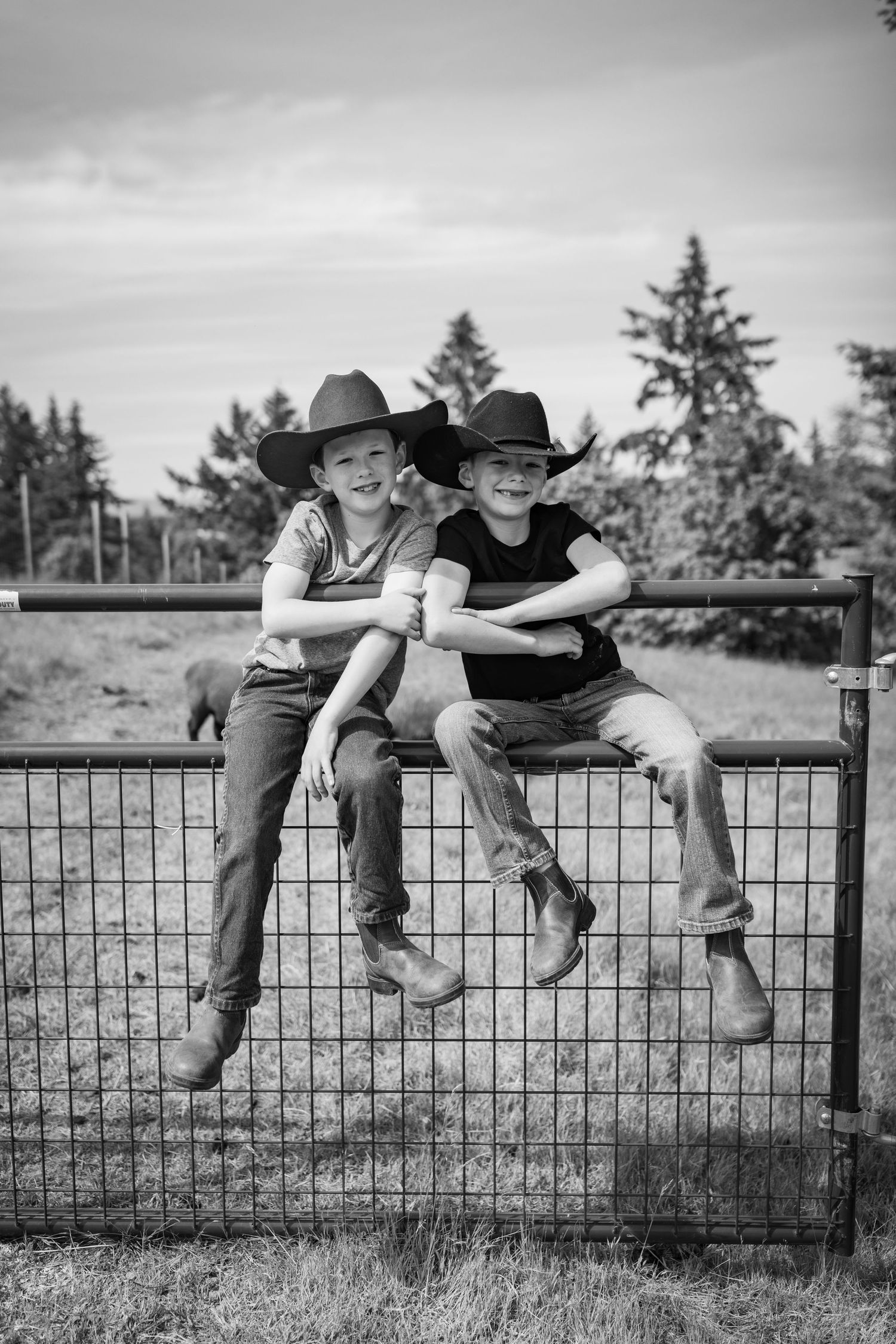 Two kids in cowboy hats sit on a metal farm gate smiling in a black and white photo.