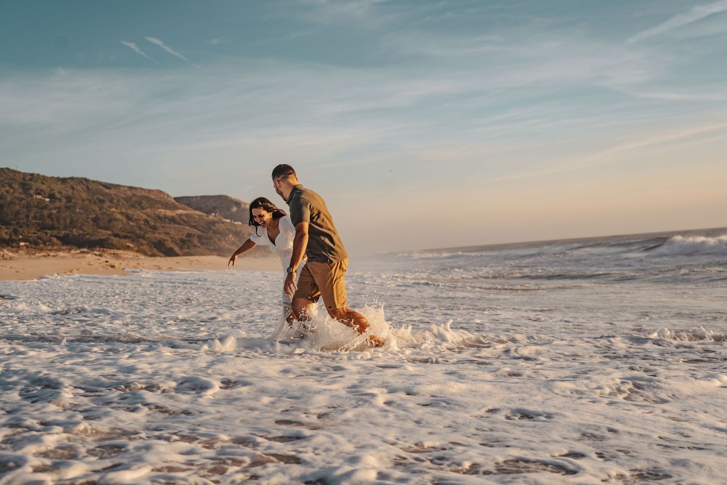 Couple running playfully on beach at sunset with waves crashing behind them.