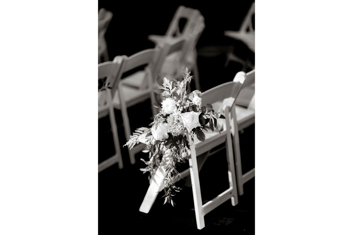 Black and white photograph of floral decorations attached to a folding chair at a wedding ceremony.