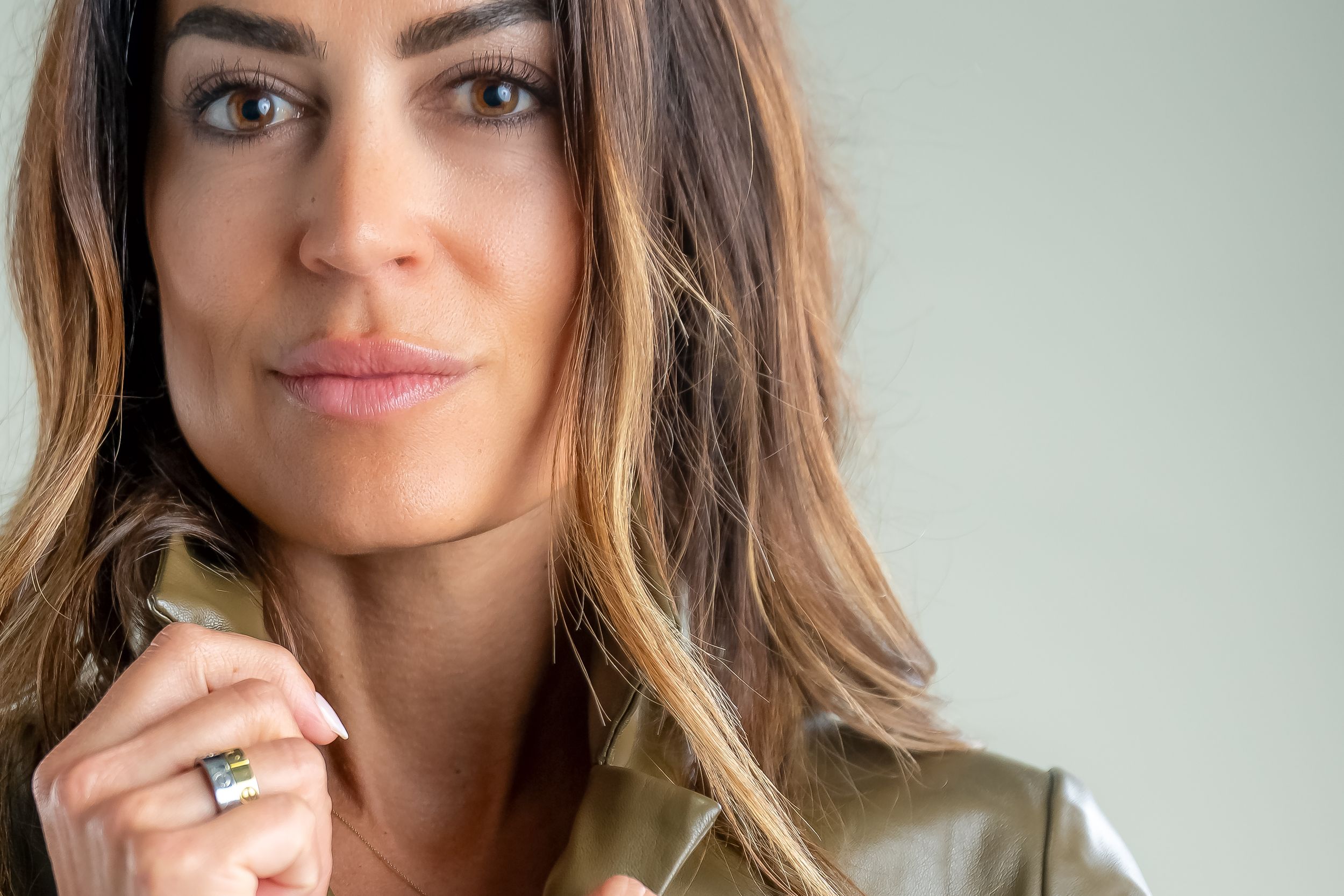 Woman with wavy brown hair wearing olive green top smiles warmly at camera in natural lighting.