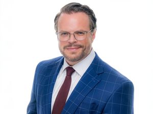 A person in a blue windowpane suit and burgundy tie smiles while wearing glasses against a light background.