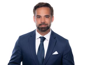 Professional headshot of a man in a navy blue suit and tie against a light background.
