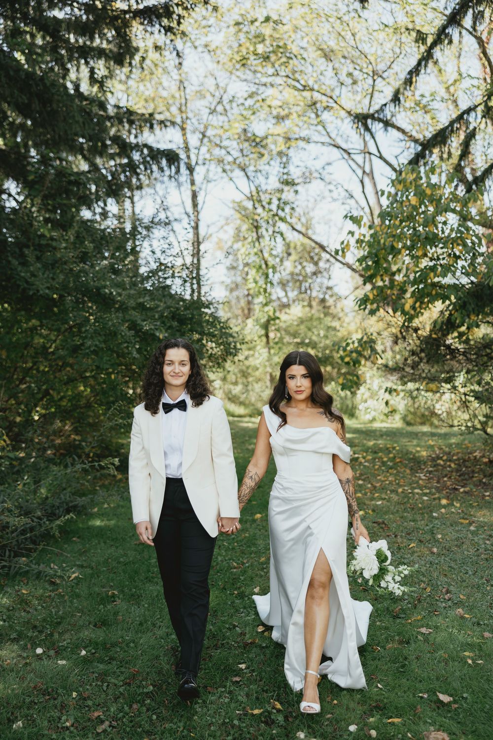 Two people in white outfits walk together through a lush green outdoor garden setting on a sunny day.