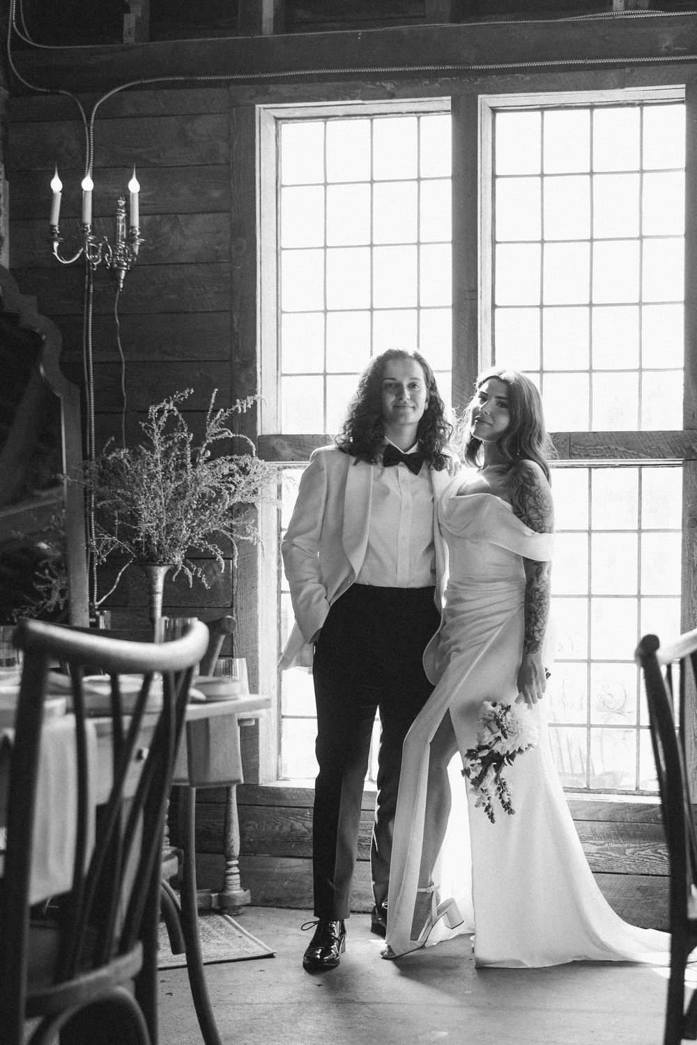 A black and white wedding photo of a couple in a rustic cabin interior with large windows and a candelabra.