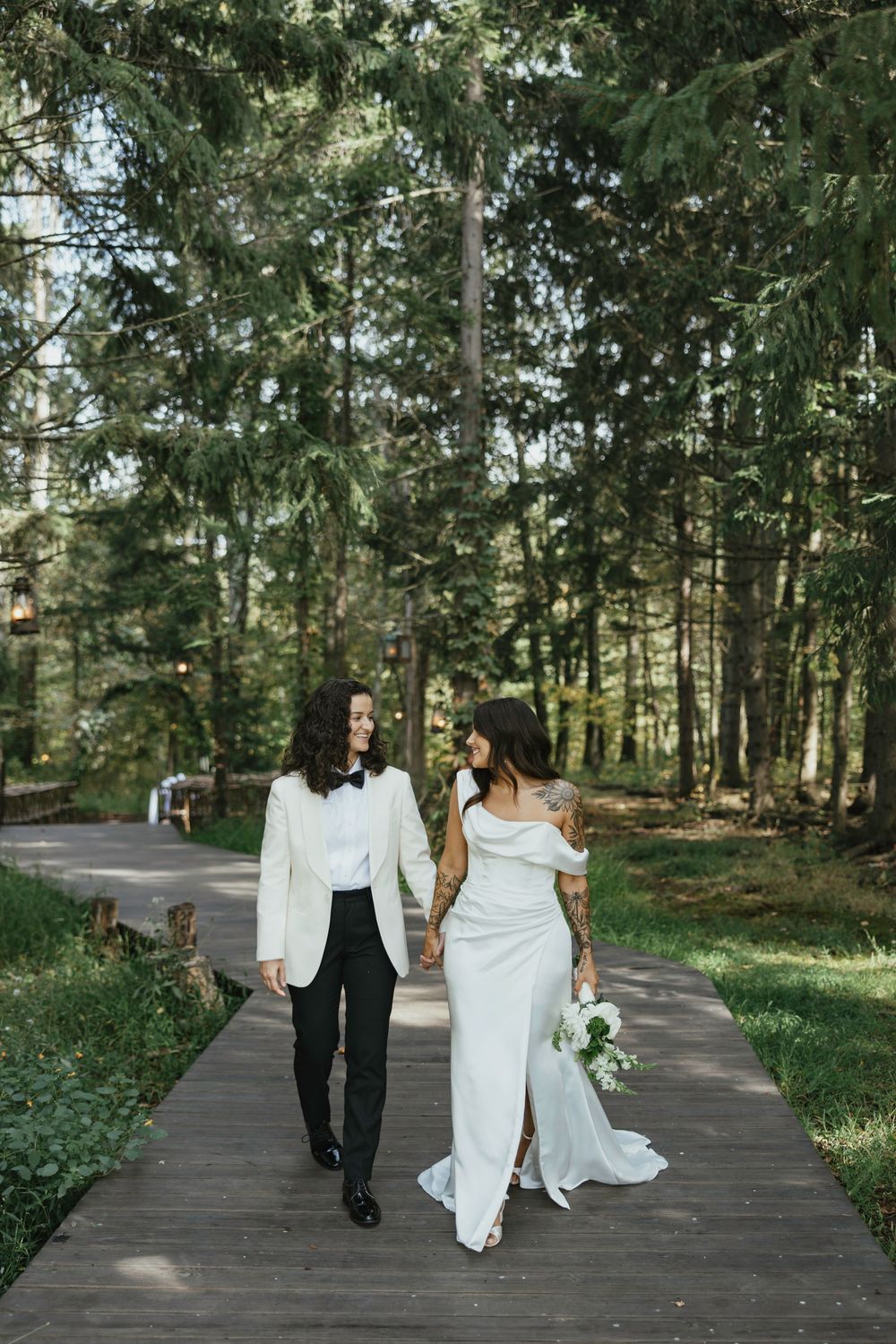 A couple in wedding attire walks together through a serene forest path lined with tall evergreen trees.