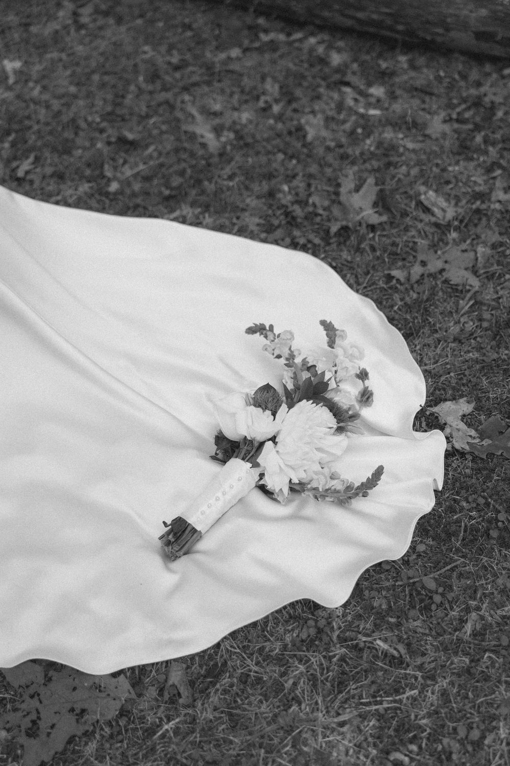 A white wedding dress veil with floral details trails across dark grass in black and white photography.