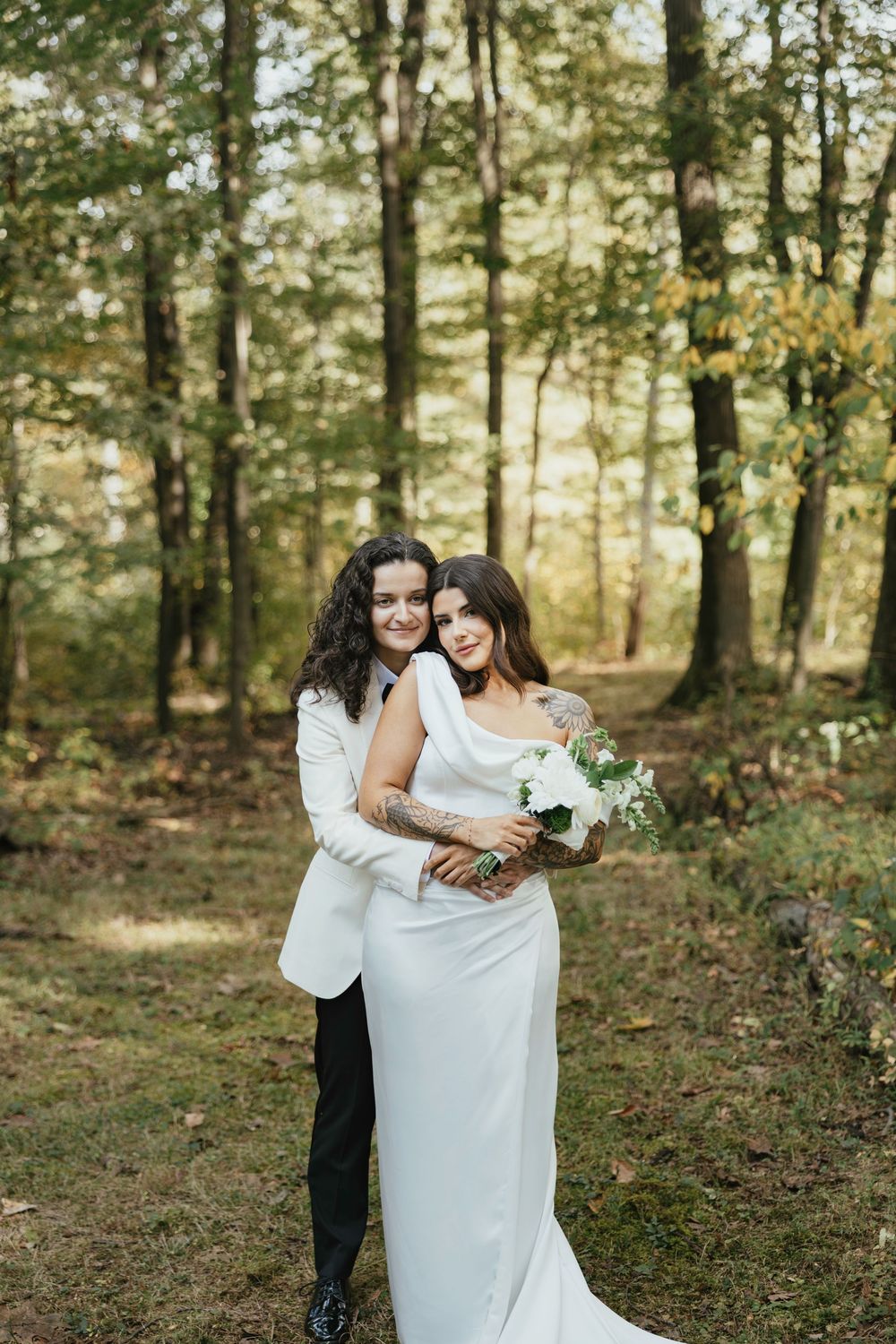 A loving couple in wedding attire embrace in a sunlit forest setting while holding a white floral bouquet.