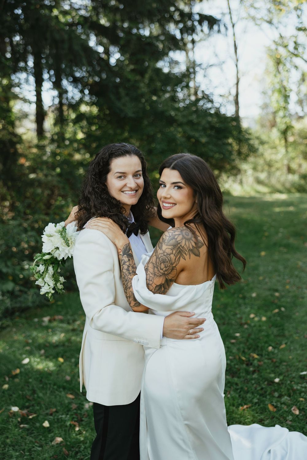 Two people in white outfits share an intimate embrace while laughing together in a sunny outdoor setting.