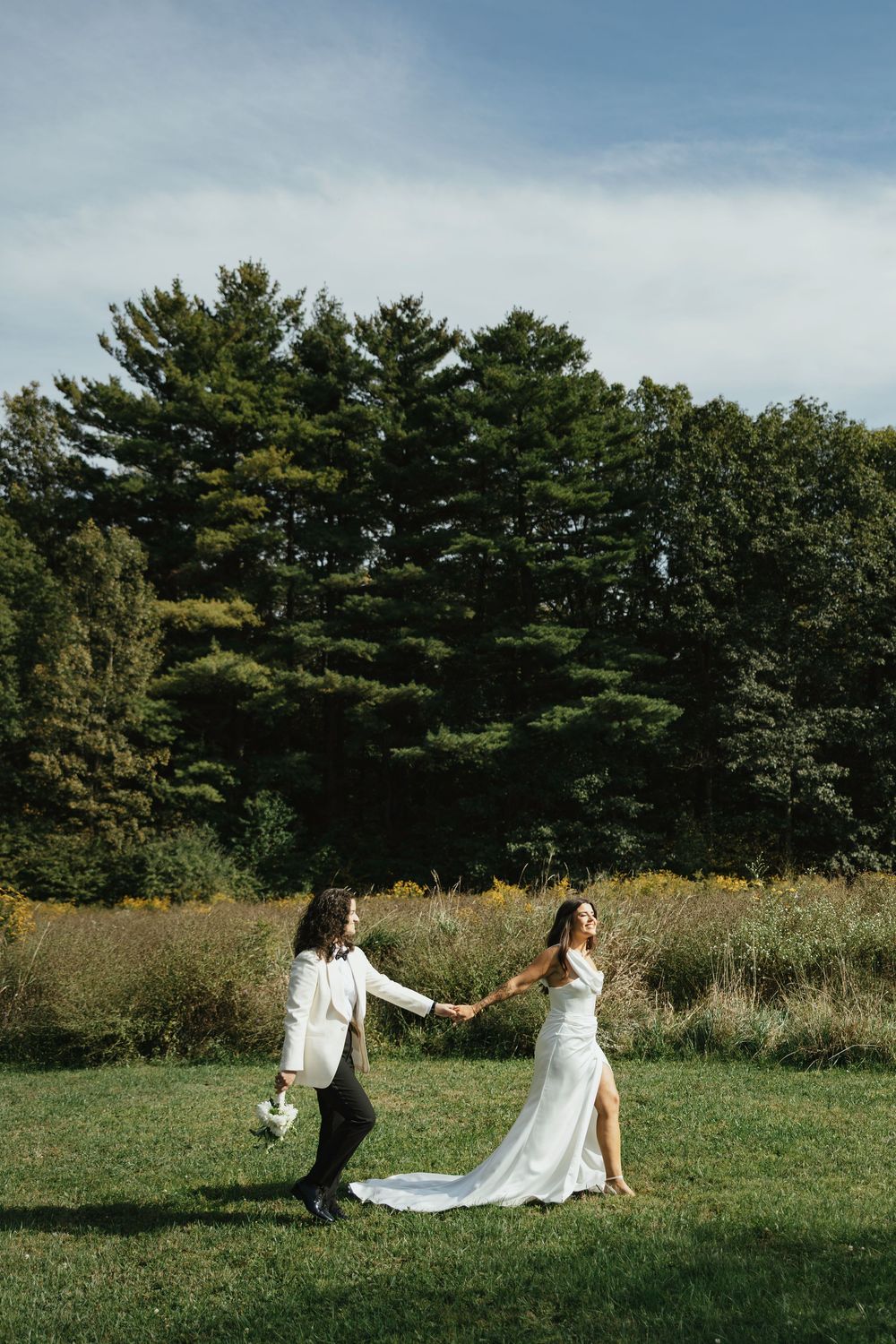 Wedding couple playfully runs and spins across a grassy field with evergreen trees in the background.