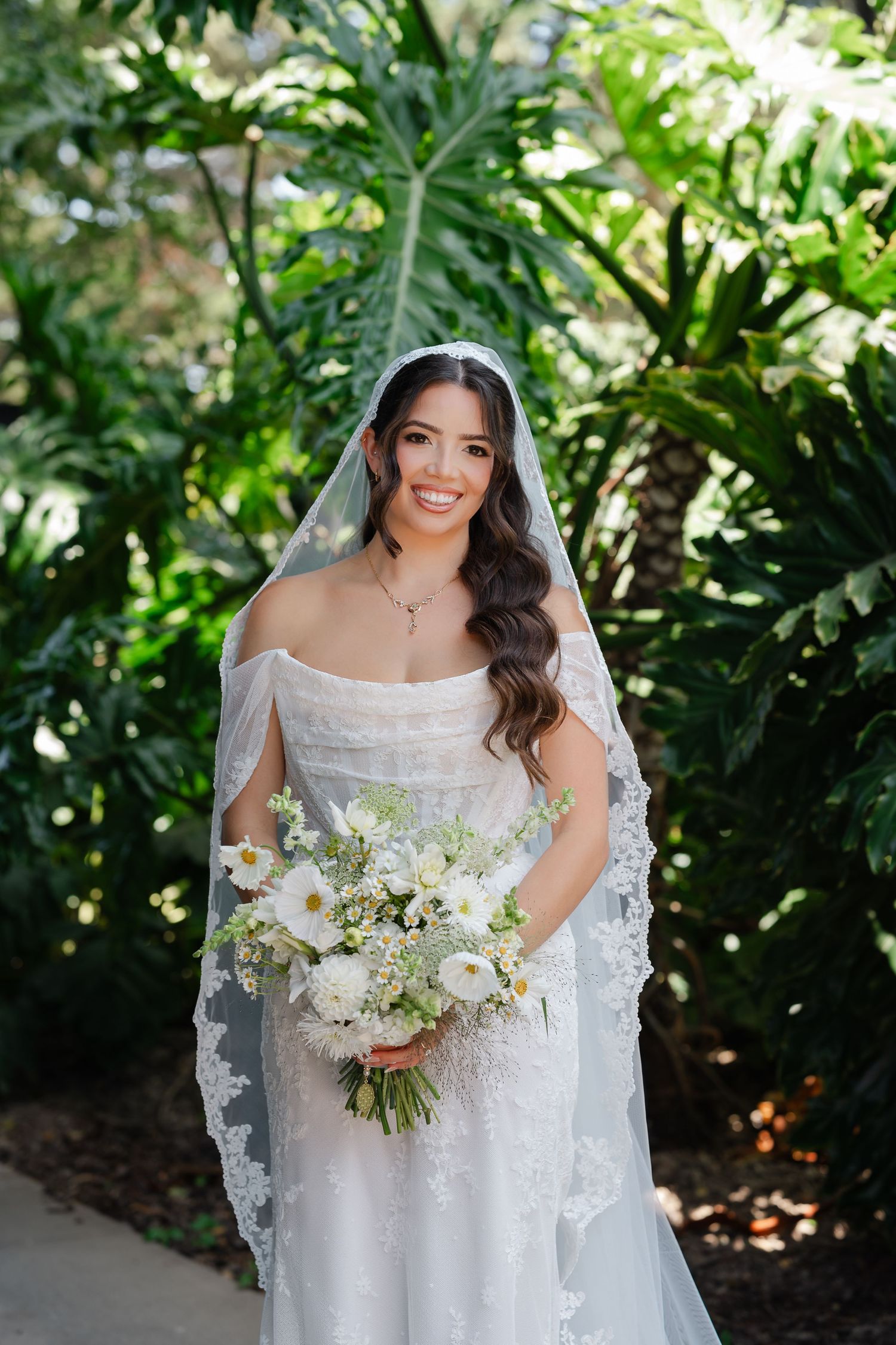 A bride wearing a white lace wedding dress and veil poses outdoors with greenery in the background.