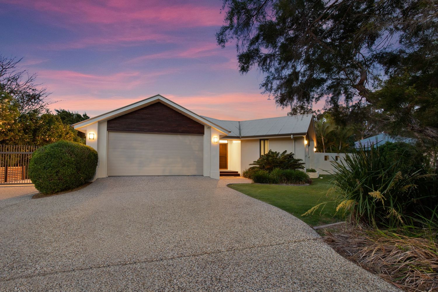 Neat single storey brick home with double garage, manicured garden and gravel driveway at sunset.