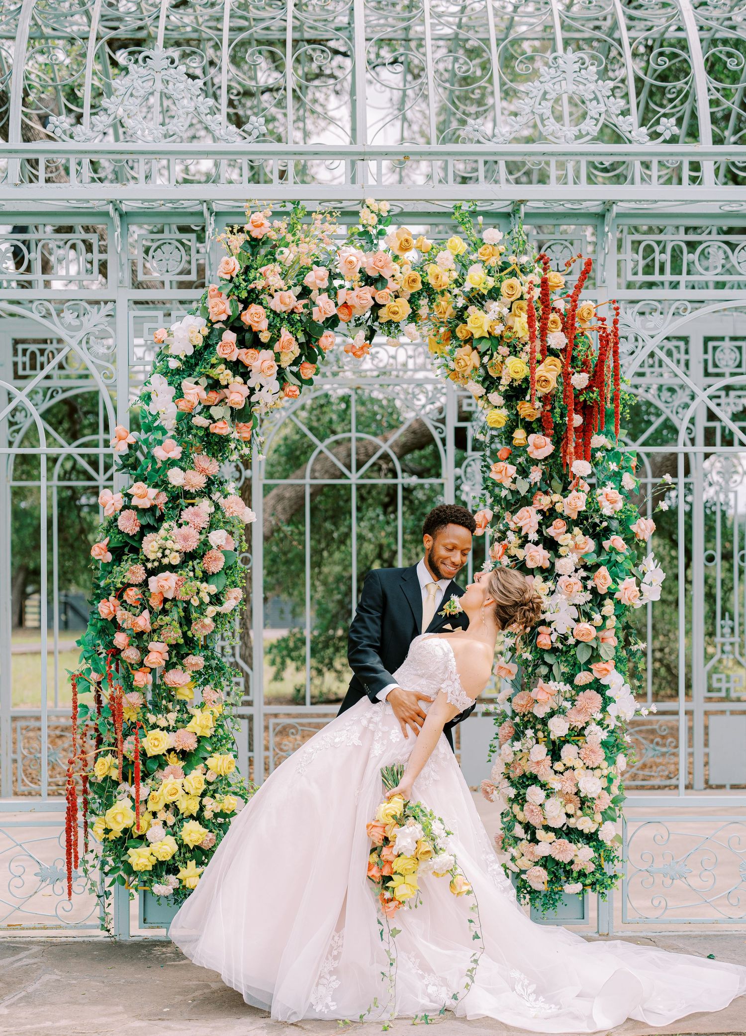 Romantic moment under floral arch with peach and yellow roses against greenhouse backdrop.