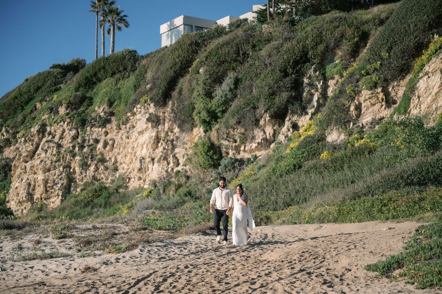 Couple walking along scenic coastal cliffs with palm trees and white houses perched above in Southern California.