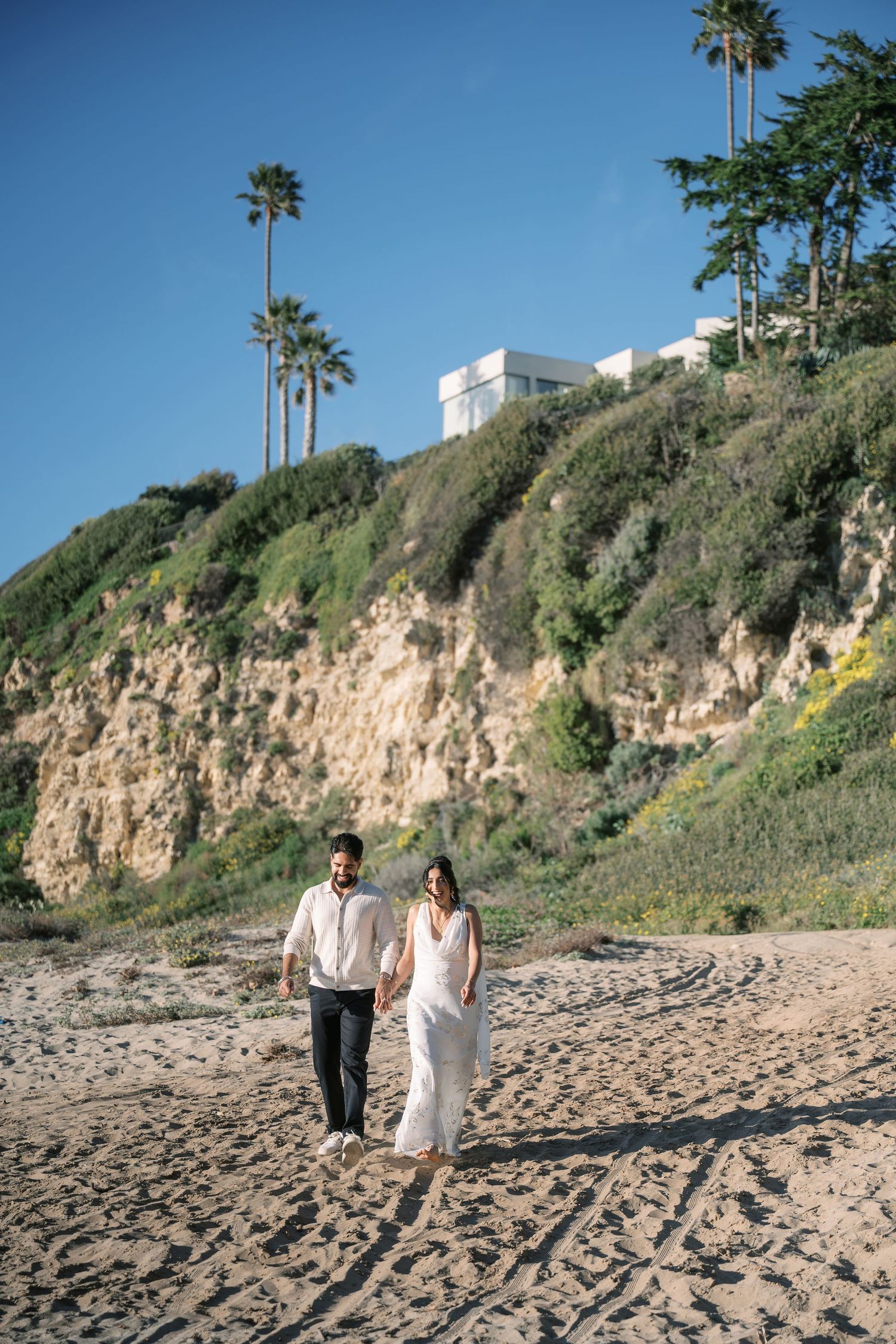 A couple walking along a scenic beach with rocky cliffs and palm trees in Southern California.