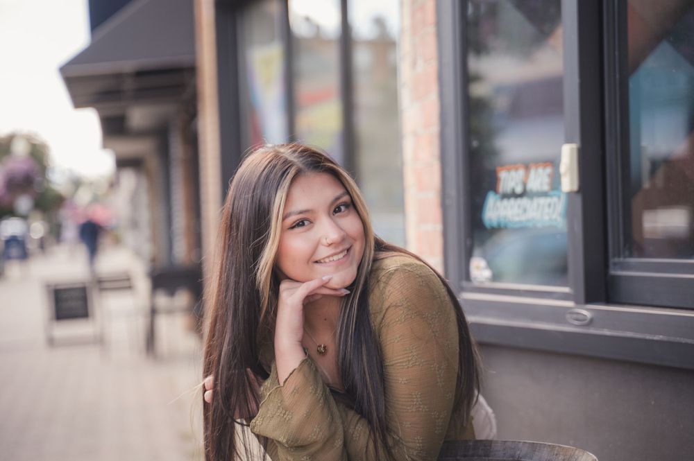 Portrait photo of someone in a brown sweater leaning against a storefront window with a soft smile.
