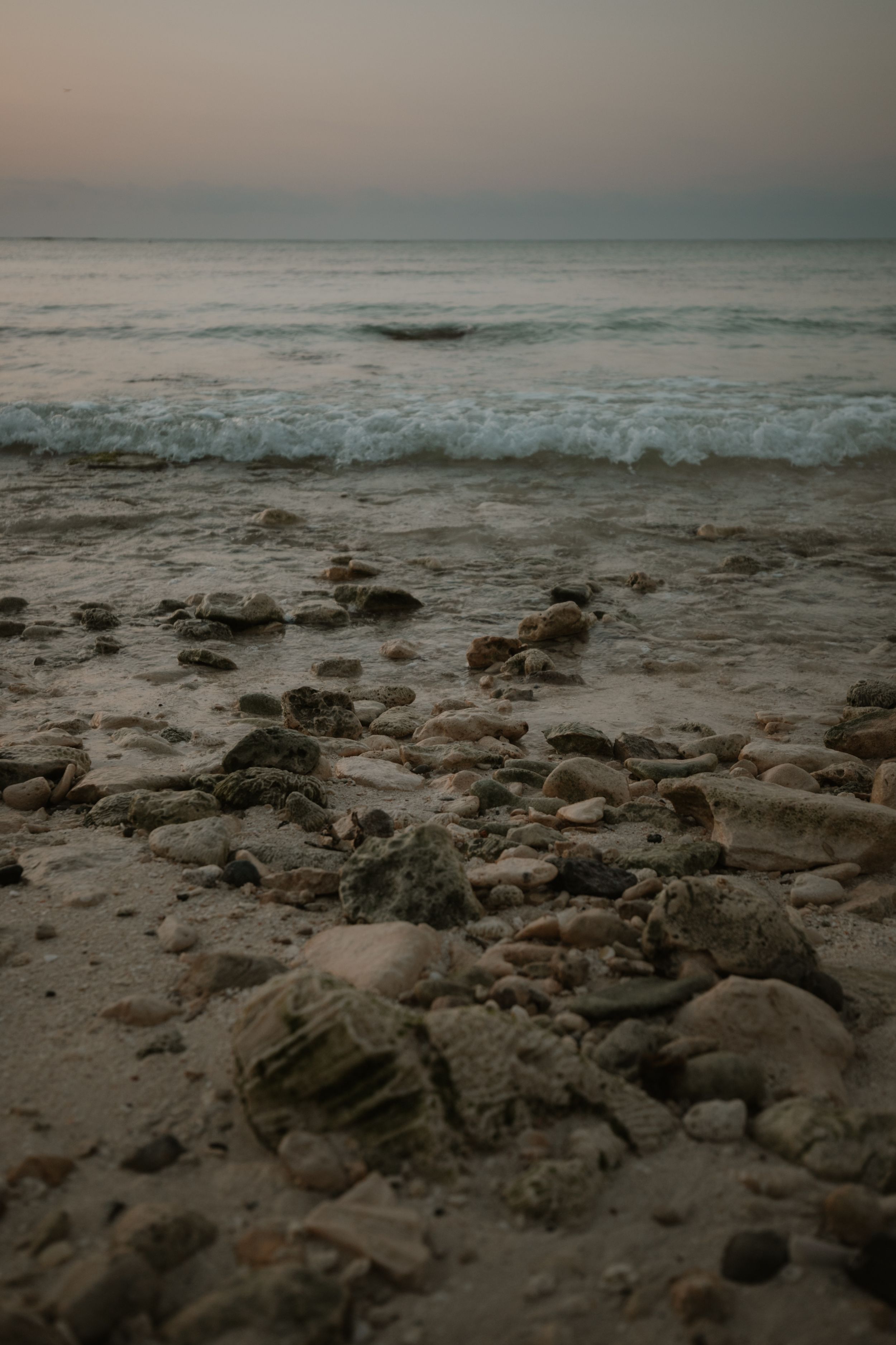 Rocky beach shoreline with gentle waves rolling in during a misty sunset over the ocean.