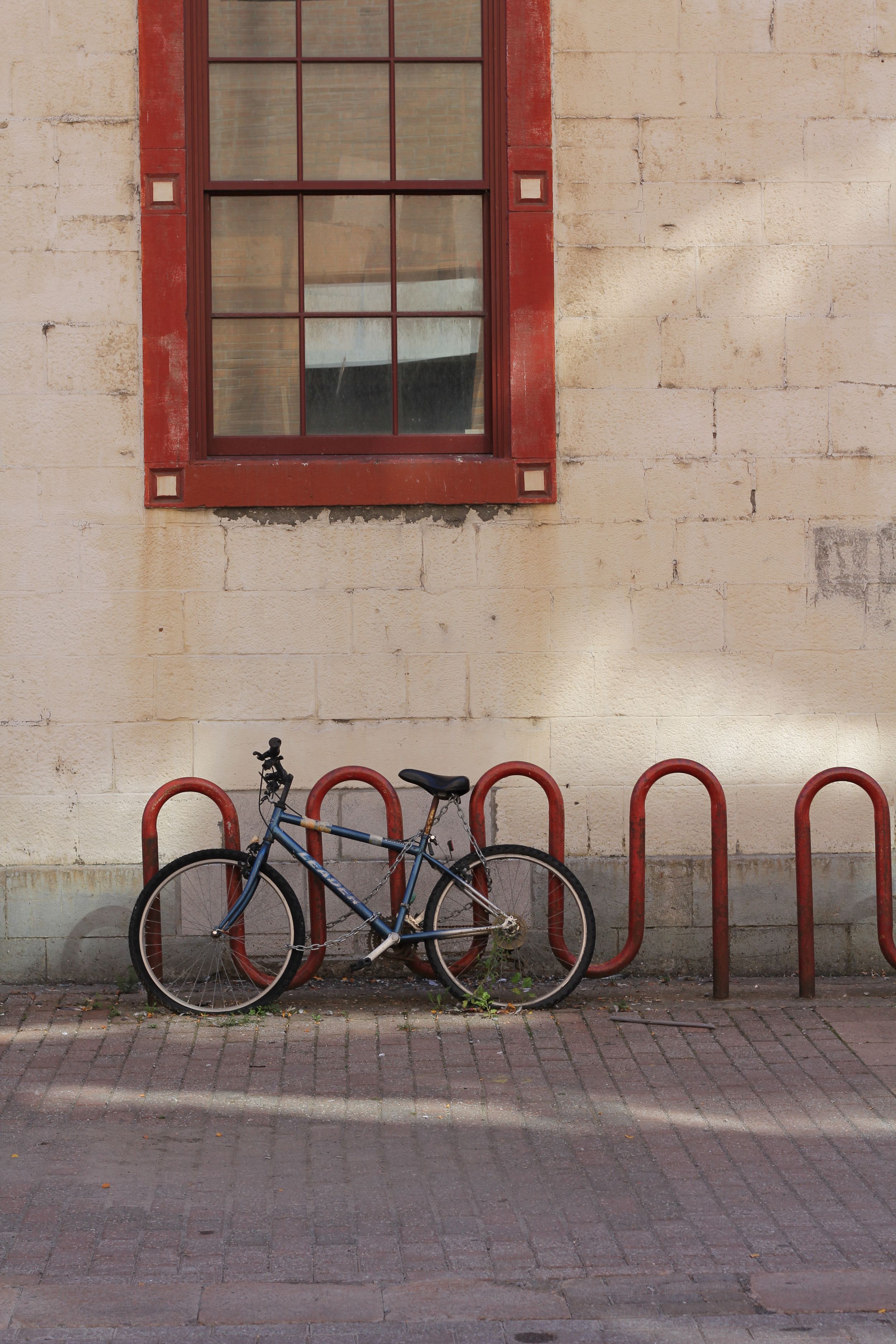 Lone bicycle parked against red bike racks beneath a window on beige building wall.