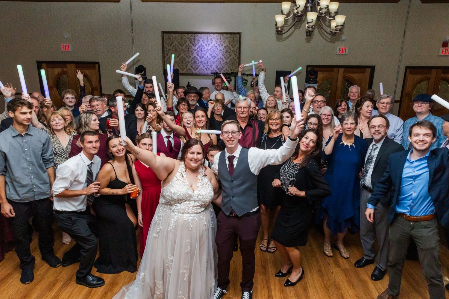 Large group of wedding guests celebrate with raised glow sticks on dance floor.