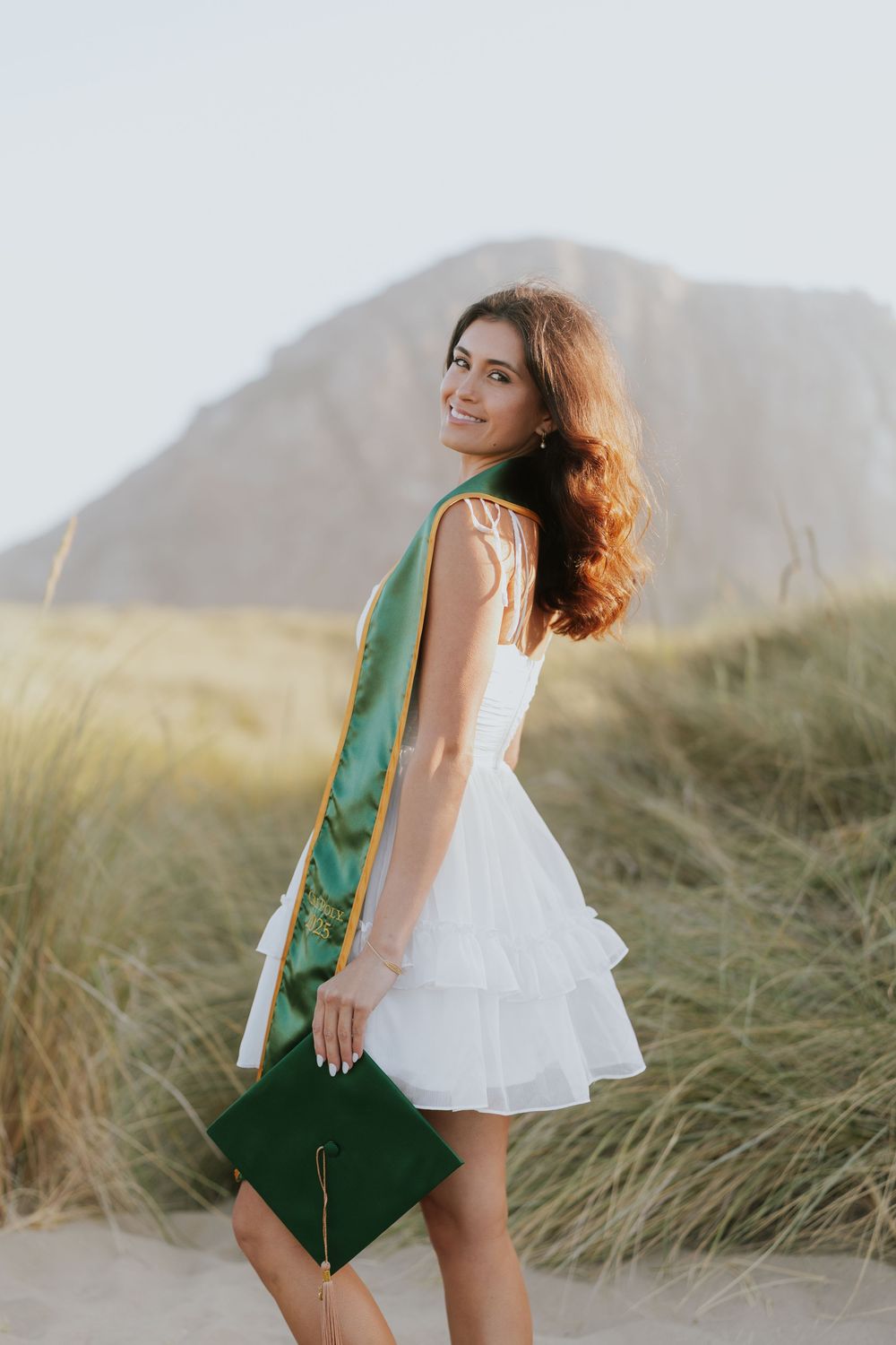 Person in white summer dress and emerald green velvet blazer poses playfully in tall beach grass at sunset.