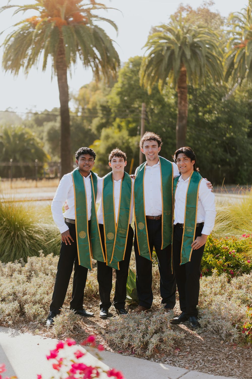Four graduates wearing green graduation stoles pose together outdoors with palm trees in the background on a sunny day.