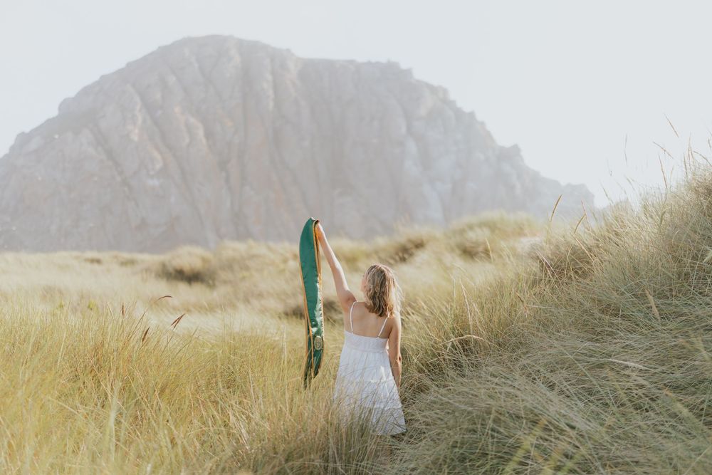 A person in a white dress stands in tall beach grass holding a colorful flag with a mountain in the background at sunset.