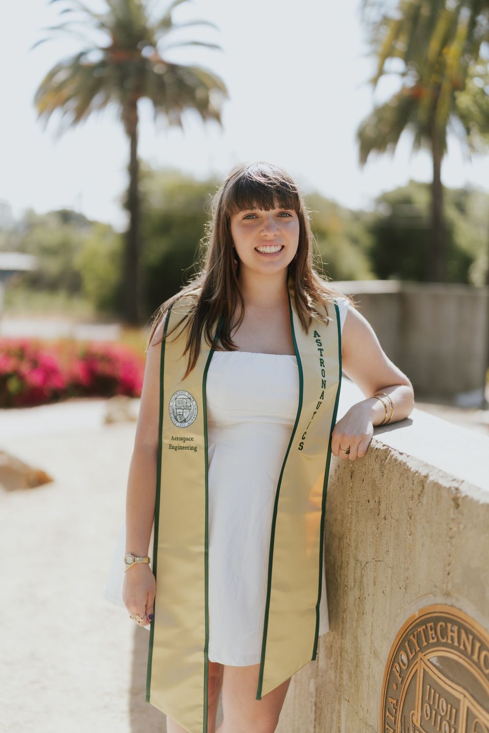 Graduate wearing a gold graduation stole and white dress poses outdoors with palm trees and flowers in the background.