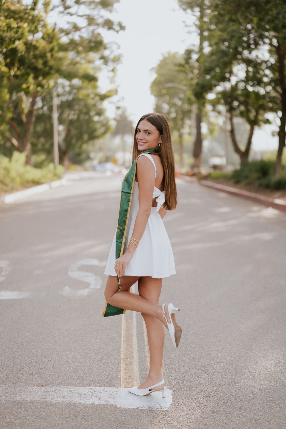Young woman in white mini dress and heels poses on a tree-lined road, smiling over her shoulder.