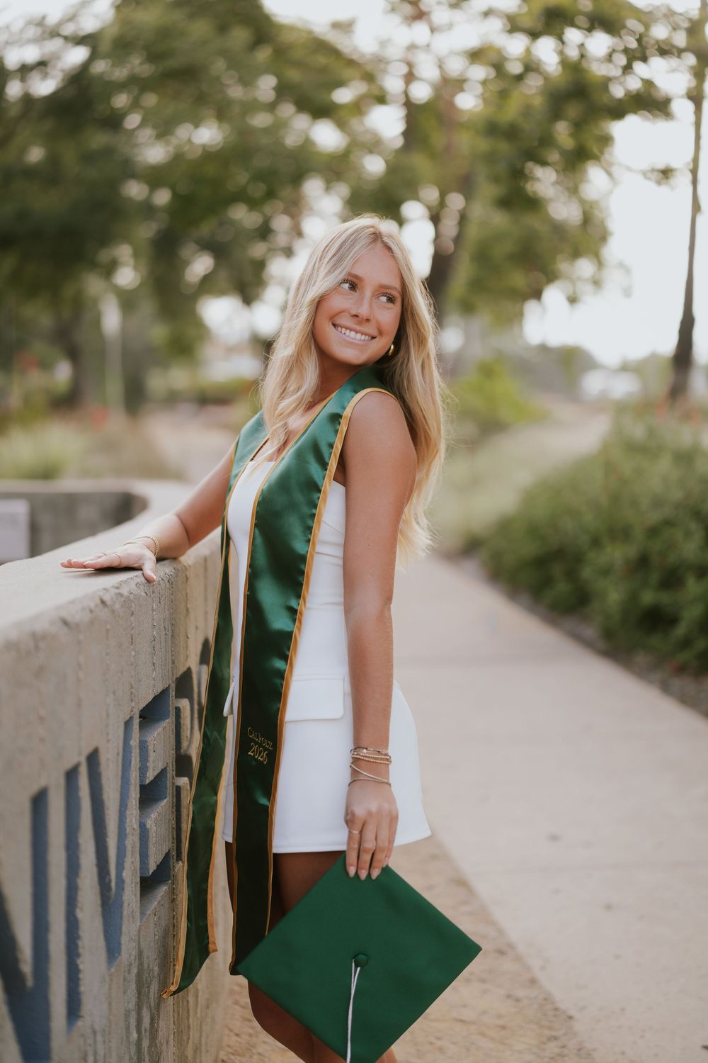 Smiling blonde woman in white dress with green graduation cap and sash poses outdoors on a sunny day.