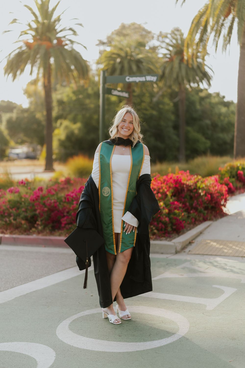Graduate in black cap and gown with green sash poses playfully on campus sidewalk with palm trees and flowers.