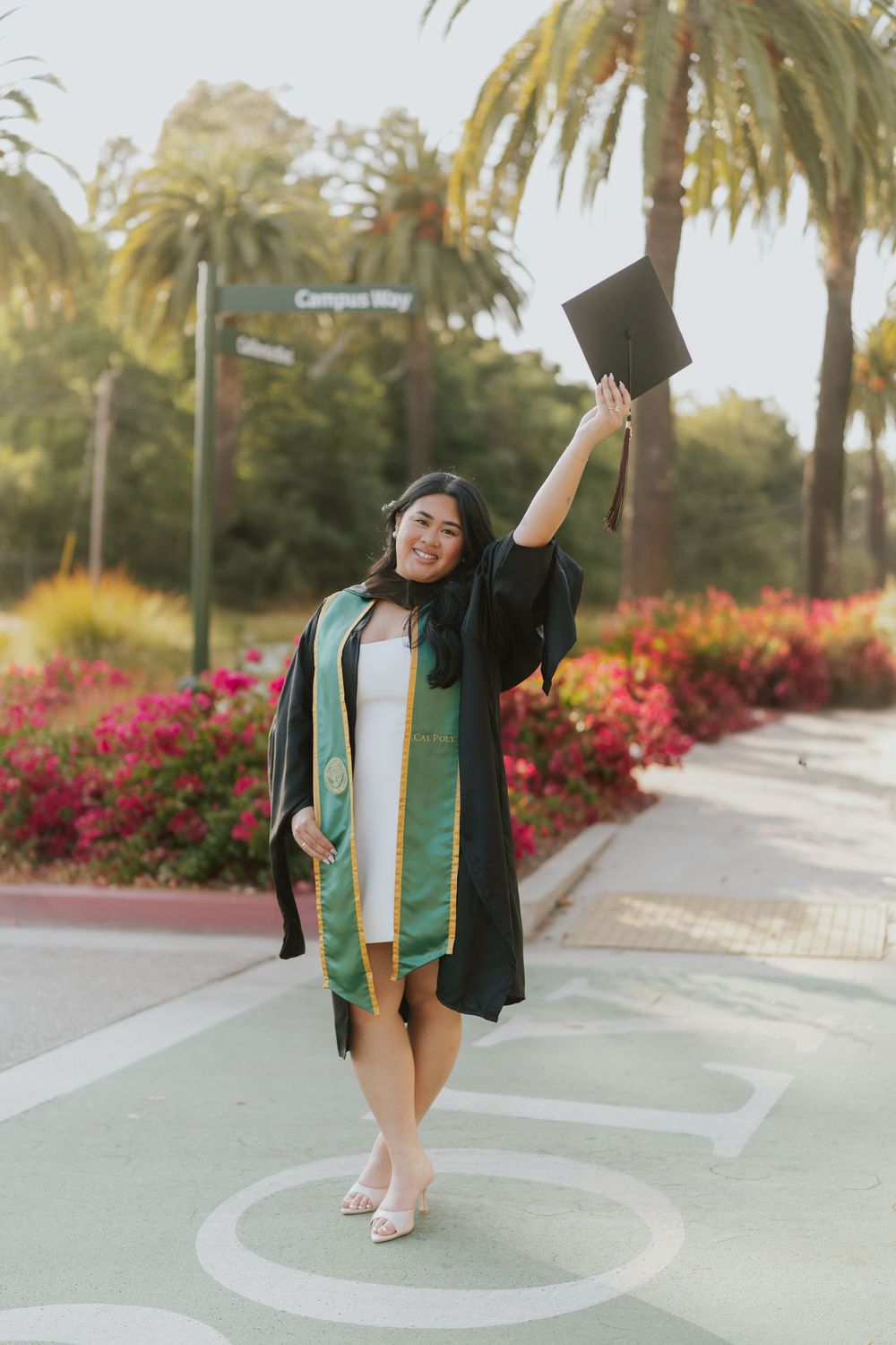 Joyful graduate in black academic regalia dances on campus pathway surrounded by palm trees and blooming flowers.