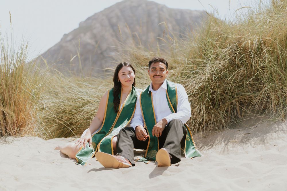 Couple in green and white outfits sitting together on sandy dunes with mountain landscape in background.