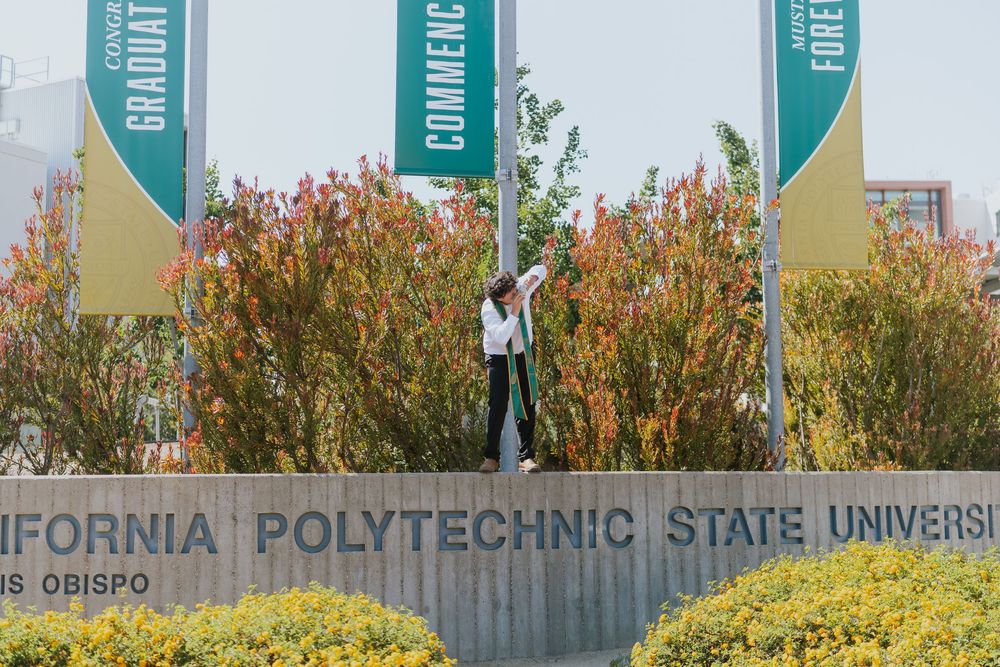 A graduate celebrating near the California Polytechnic State University entrance sign surrounded by colorful landscaping.