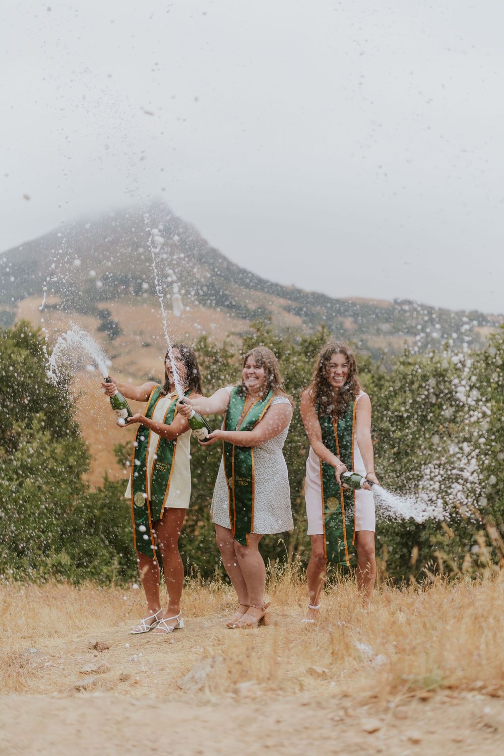 Friends celebrating with champagne sprays on a mountainside trail during golden hour.