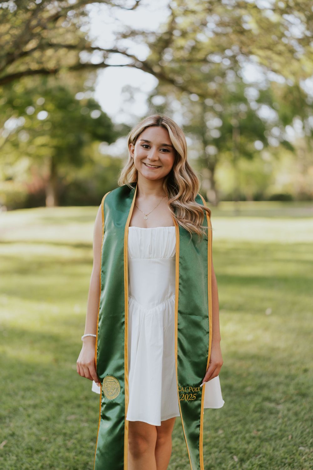 Graduate wearing green academic stole and white dress stands smiling on sunny lawn during outdoor graduation photoshoot.