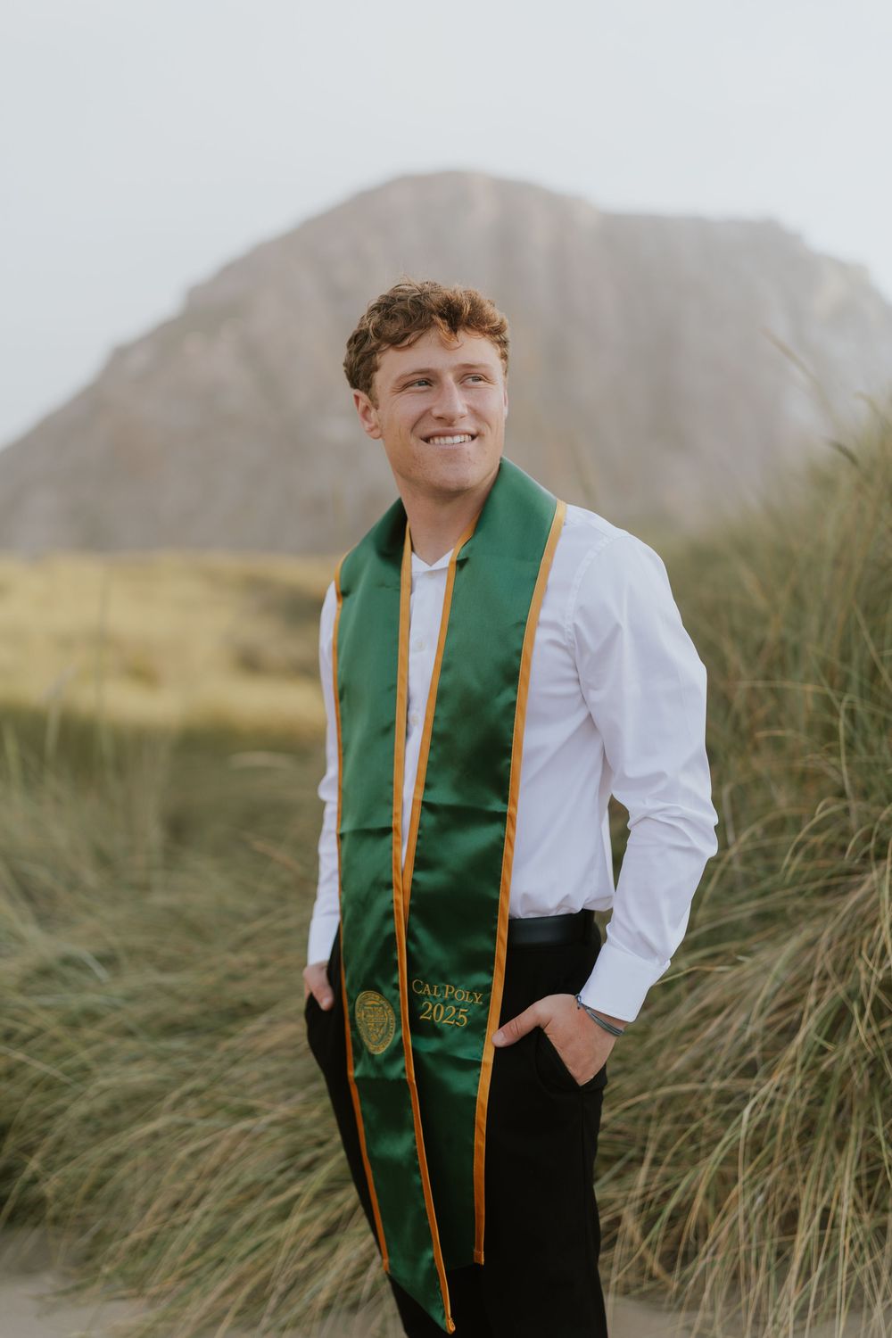 Graduate wearing green and gold graduation stole stands in tall grass with mountain in background on sunny day.