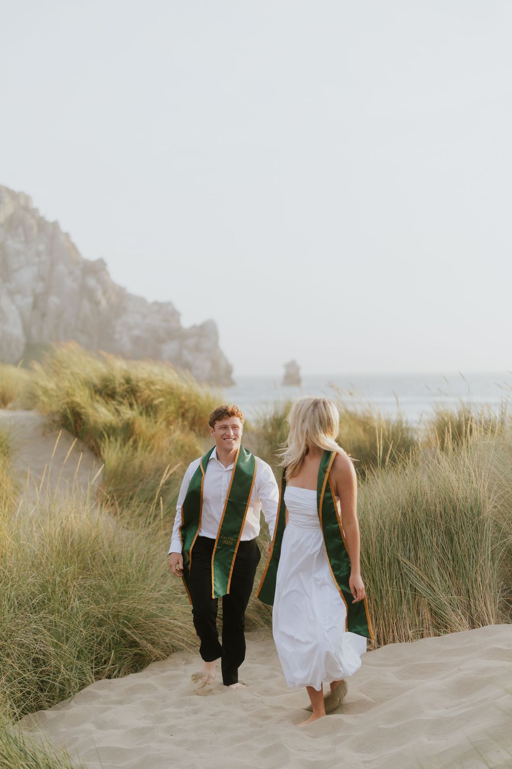 A couple walks barefoot together through coastal sand dunes with tall grass and cliffs in the background.