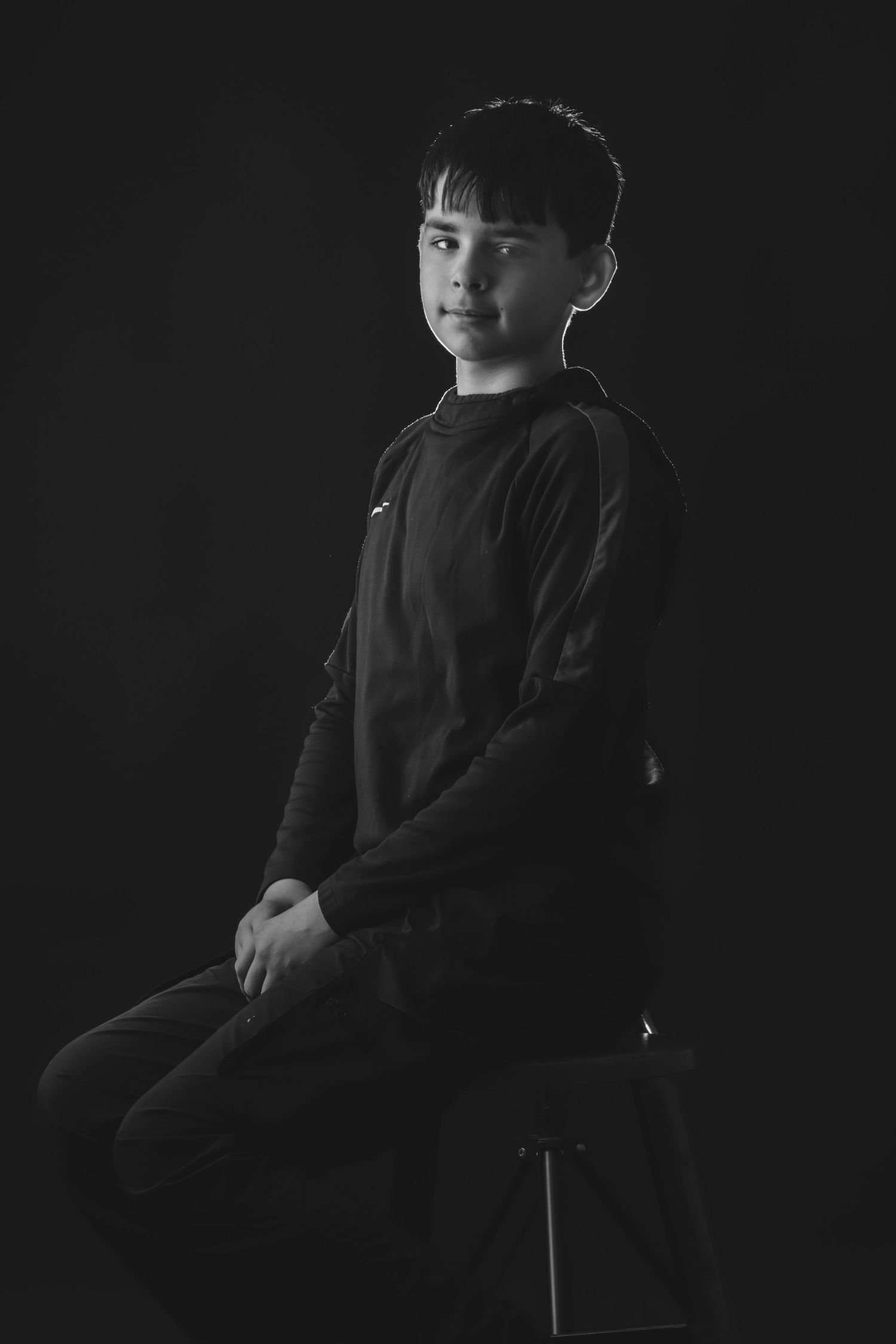 A dramatic black and white studio portrait of a person seated on a stool.