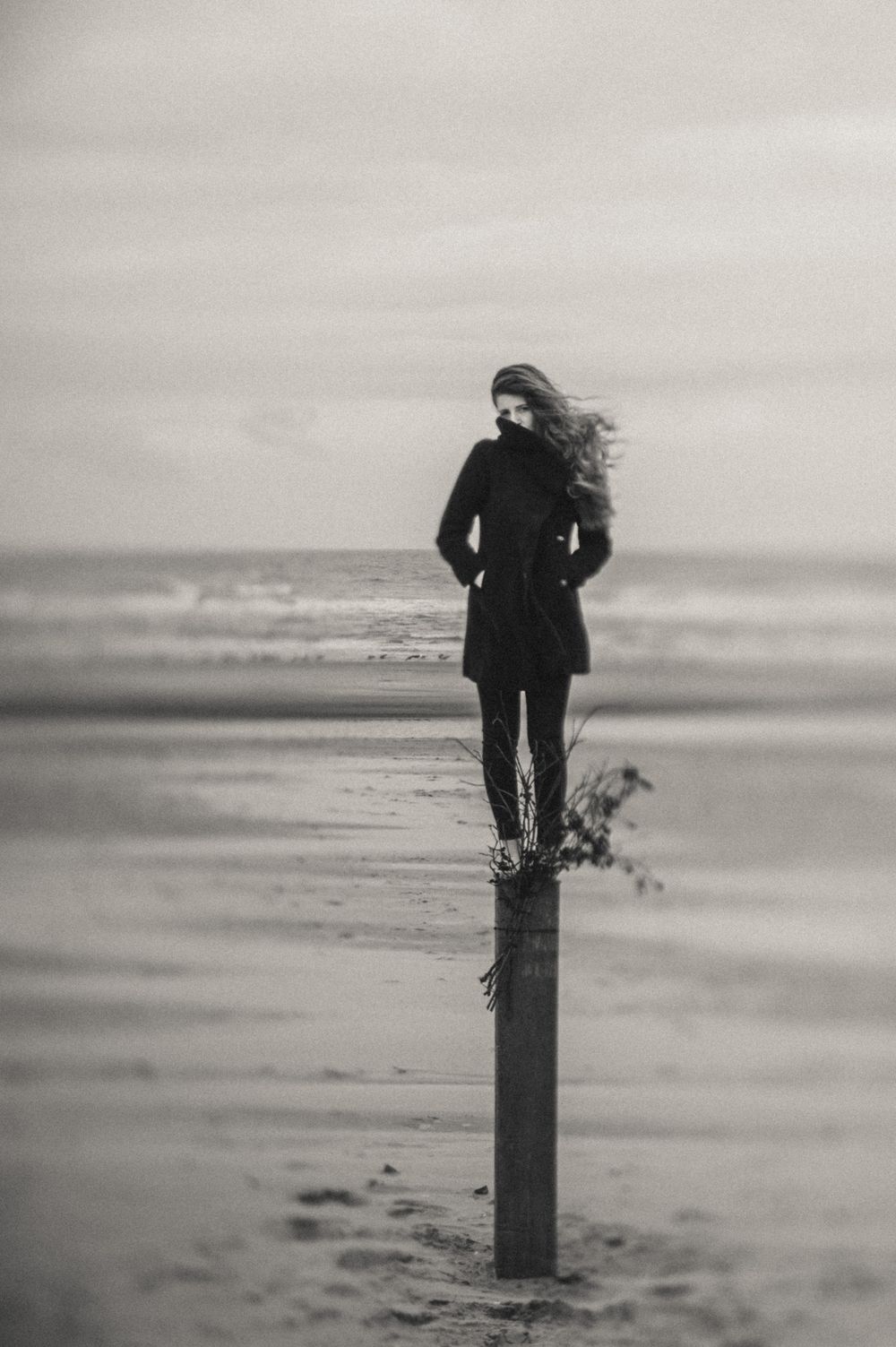 Moody black and white photograph of someone perched on a post at a misty beach.