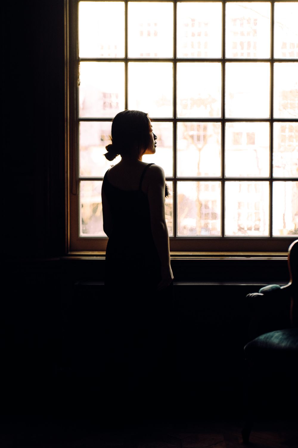 Silhouetted figure sits by window in dark room creating moody atmospheric scene.